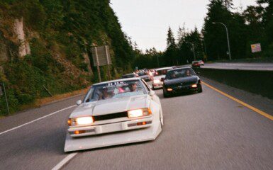 Vintage cars cruising on a scenic highway lined with trees, showcasing retro automotive culture.