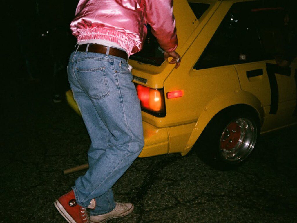 Person in a pink jacket and jeans leaning on a yellow car with a raised hatch at night.