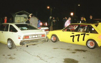 Group of people gathered around vintage cars at night, featuring a yellow car with racing number 777.