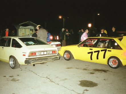 Group of people gathered around vintage cars at night, featuring a yellow car with racing number 777.