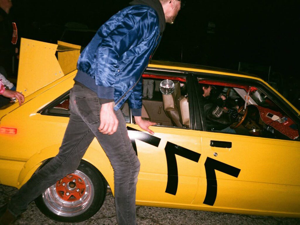 Man opening yellow car door with black decals at night street event.
