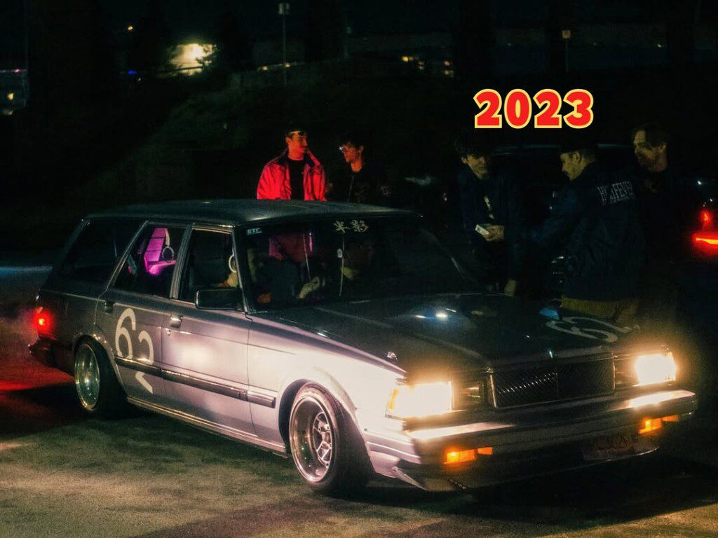 Vintage sedan car at a night gathering with people around it, illuminated by headlights, 2023.