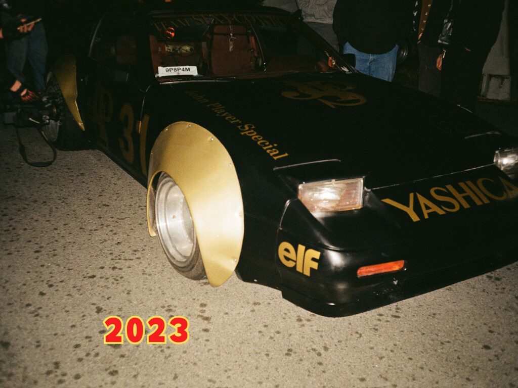 Vintage racing car with Yashica and Elf branding, parked with 2023 overlay. Dark surroundings, people nearby.