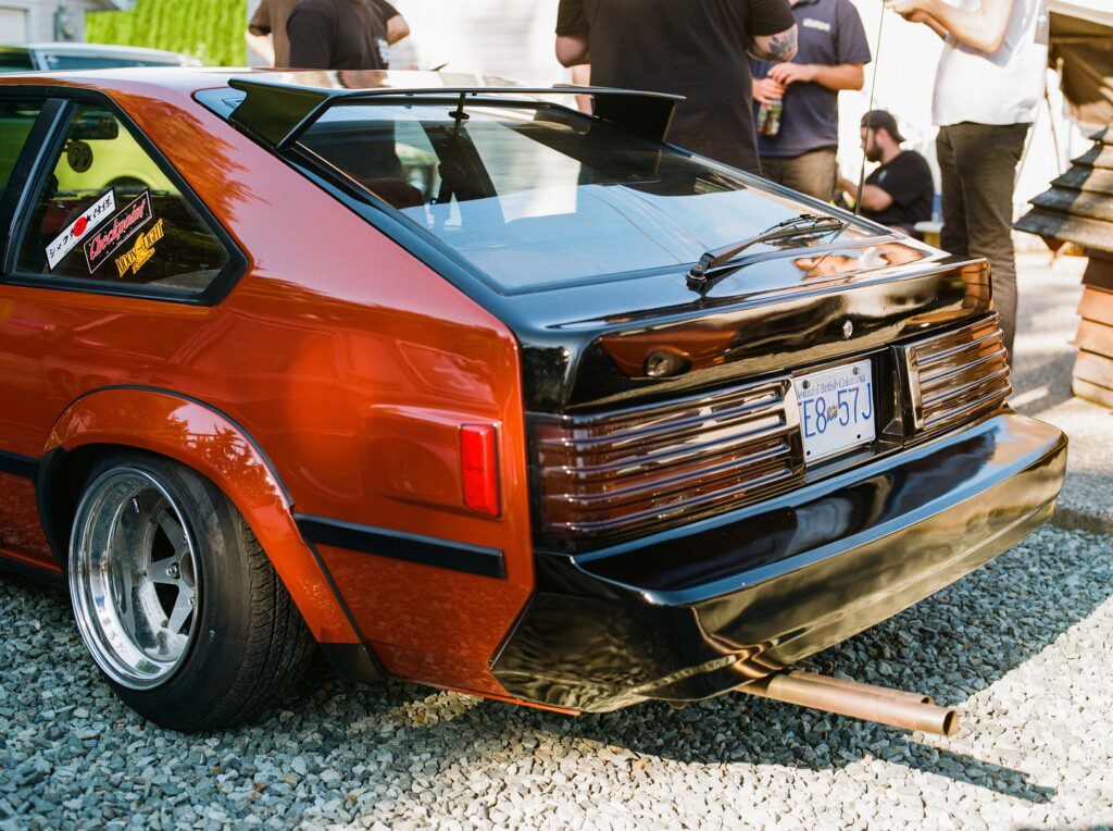 Vintage red and black sports car with custom rims and spoiler, parked on gravel with people in the background.