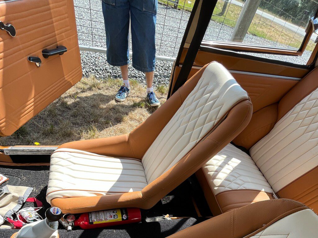 Vintage car interior with tan and cream seats, door open, fire extinguisher on floor, person standing outside.
