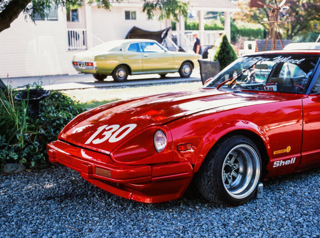 Shiny red race car number 130 parked on gravel with a vintage car in the background.