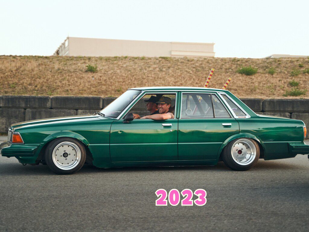 Vintage green car with a driver on the road, 2023 in bold pink text below.