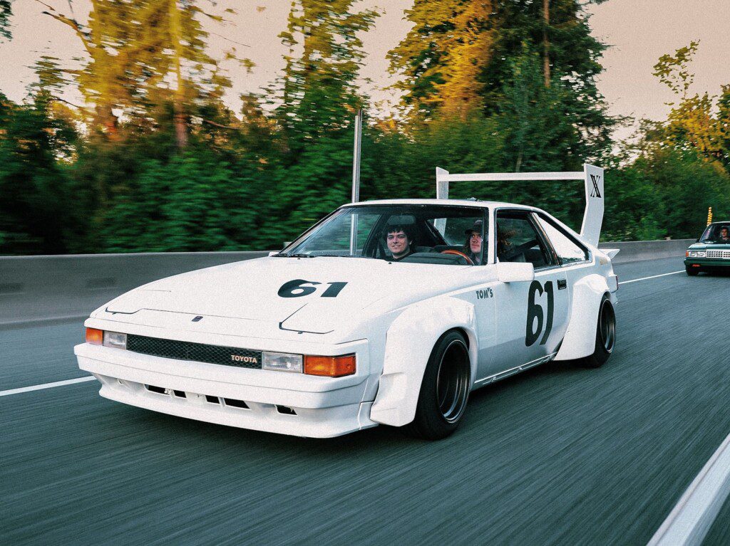 Vintage white race car with number 61 driving on a highway, surrounded by greenery in the background.