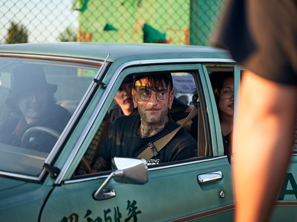 Man with tattoos driving vintage car with passengers, smiling through open window, sunny day, fence in the background.