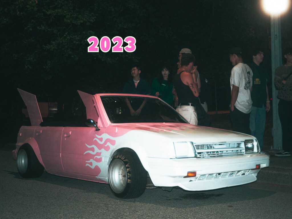 Customized white and pink car with winged doors at night, group of people nearby, 2023.