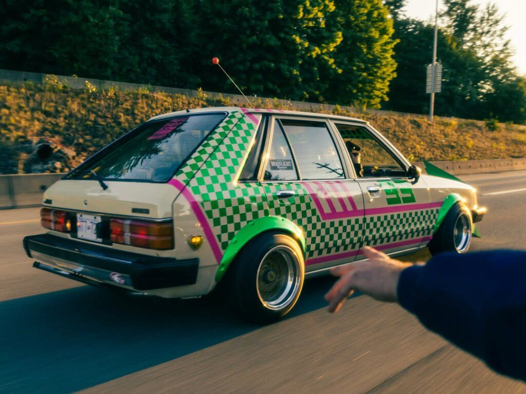 Colorful retro car with green checkerboard design cruising on highway at sunset, hand pointing from foreground.
