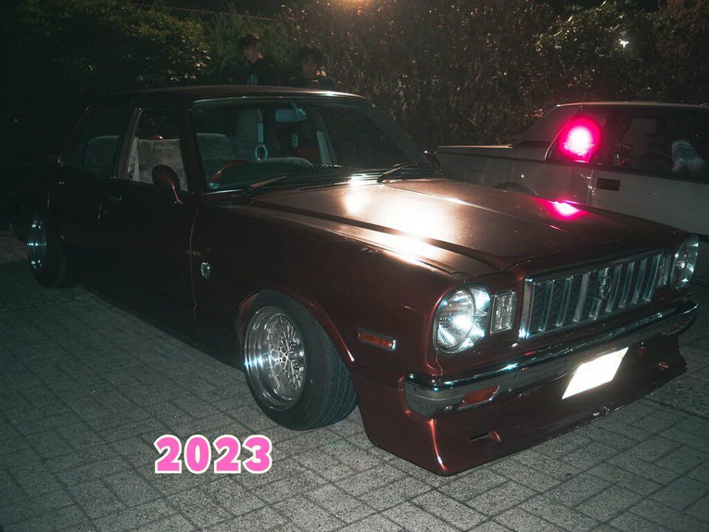 Vintage maroon car parked at night, illuminated with glowing headlights, 2023 in pink text in foreground.