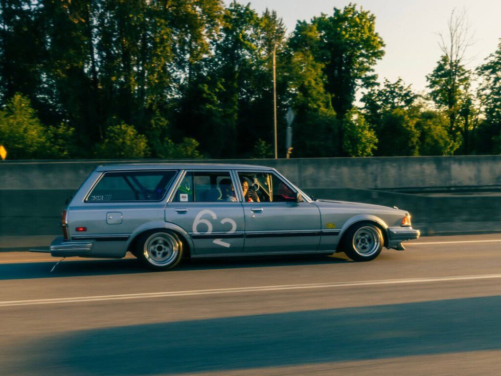 Vintage silver station wagon with number 62 racing on the highway amidst green trees.