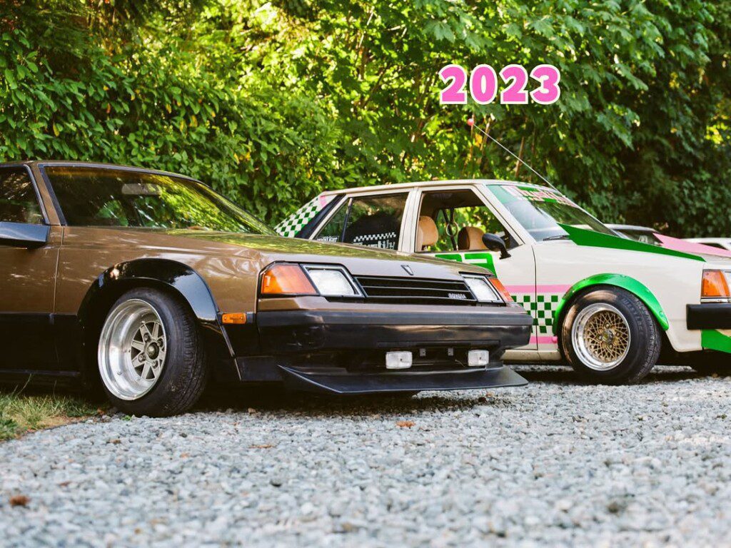 Vintage two-tone cars parked on gravel with trees in the background, marked with 2023.
